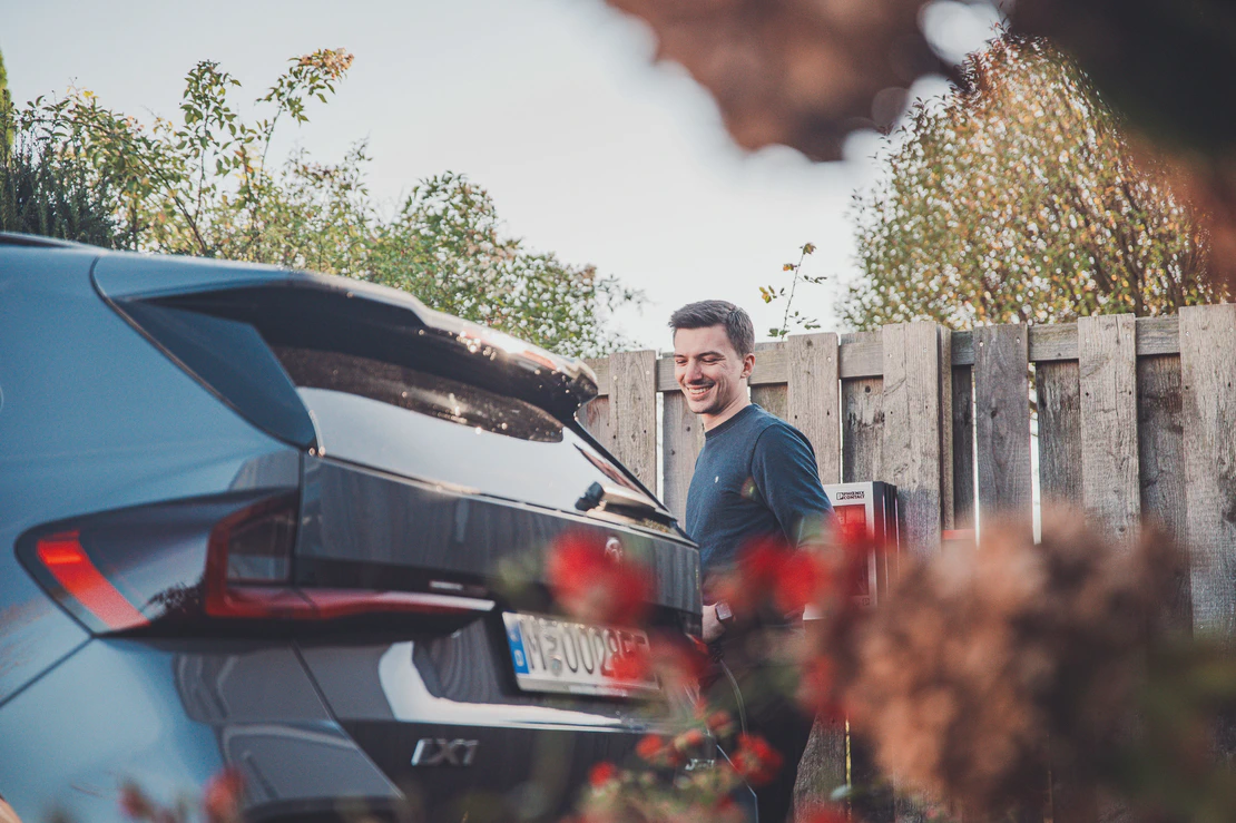 Eine Person steht neben dem Heck eines grauen Autos mit europäischem Kennzeichen. Im Hintergrund sind ein Holzzaun und etwas Grün zu sehen, im Vordergrund unscharfe Blumen. 